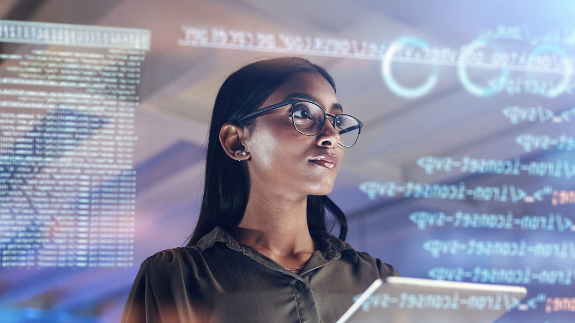 dignified mocha-skinned lady with glasses looking off axis with abstract computer code sumperimposed on large screen behind her.
