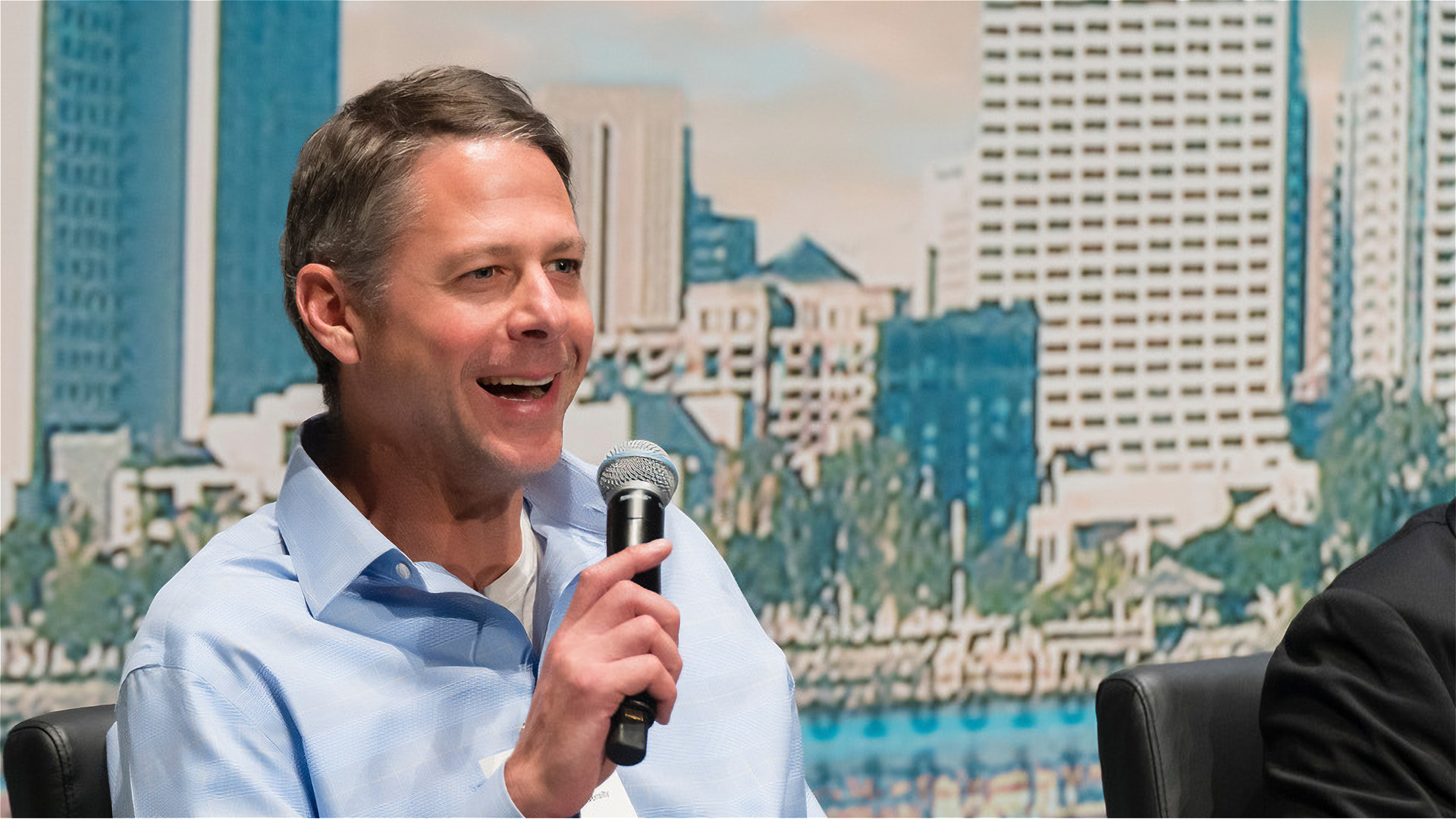Robert Beverly holding a microphone sitting on a chair and smiling as being interview on a panel.